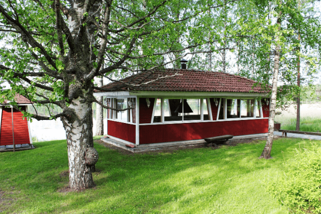 Punainen rakennus, jossa on valkoinen katto ja ikkunanpuitteet, Ympärillä on nurmikkoa ja koivuja. Taustalla näkyy järvi. Aurinko paistaa, ja maisema on kesäinen ja rauhallinen.