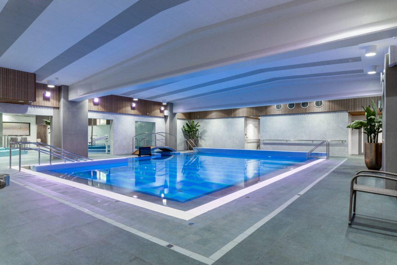 Sisäuima-allas, jossa on moderni ja tyylikäs ilme. Altaan ympärillä on harmaita laattoja ja katossa on valkoisia ja harmaita paneeleja. Vesi on kirkasta ja sinistä, ja altaan reunalla on valkoisia kaiteita. Tilassa on suuria viherkasveja ruukuissa. Seinät ovat vaaleita, ja niissä on puupaneelikoristeita. Valot ovat kirkkaat ja luovat tilaan miellyttävän valaistuksen.