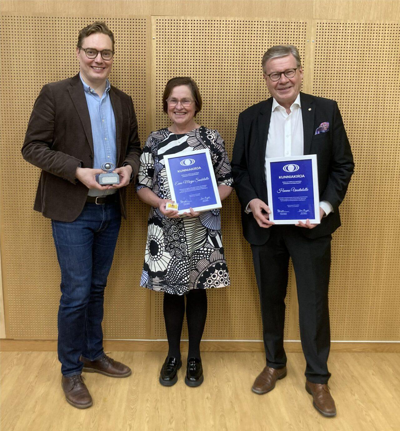 Kolme iloisesti hymyilevää henkilöä seisoo rinnakkain pidellen käsissään huomionosoituksia. Tummaan pukuun ja vaaleansiniseen kauluspaitaan pukeutunut mies pitelee kivijalustaan kiinnitettyä silmän muotoista metallilogoa. Kirjavaan mekkoon pukeutuneella naisella ja tummaan pukuun pukeutuneella miehellä on kunniakirjat, joissa on valkoiset kehykset ja valkoista tekstiä sinisellä pohjalla.