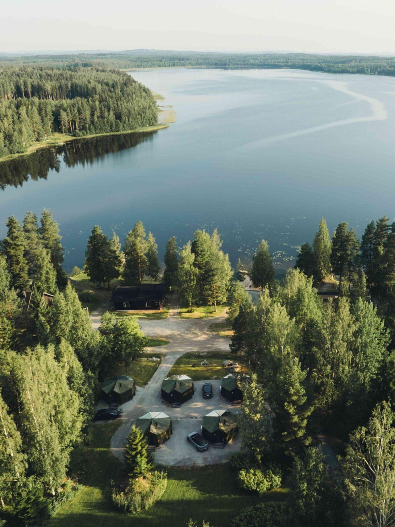 Ilmakuva järvimaisemasta ja mökkialueesta. Kuvan yläosassa on suuri tyyni järvi, jota ympäröi tiheä havu- ja sekametsä. Järven rannalla on pieniä mökkejä, joiden katot pilkistävät puiden välistä. Kuvan alaosassa näkyy viisi kuusikulmaista mökkiä sekä useita pysäköityjä autoja. Mökkien ympärillä on runsaasti vihreää luontoa ja puita. Tunnelma on rauhallinen ja luonnonläheinen, tyypillinen suomalainen järvimaisema. Sää on aurinkoinen.