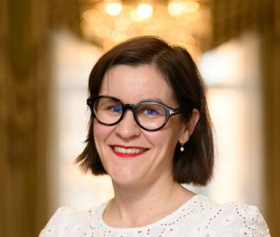 Hymyilevä nainen, jolla on lyhyet tummanruskeat hiukset ja mustasankaiset silmälasit. Hänellä on yllään valkoinen, pitsikuvioinen paita. Tausta on valaistu, epätarkka, kultainen ja lämminsävyinen.