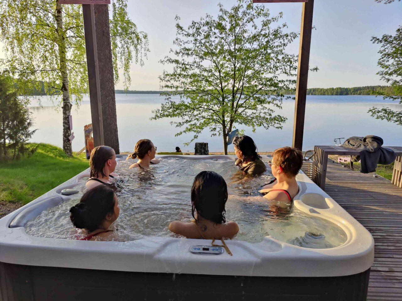 Ulkoporeallas, jossa istuu kuusi ihmistä. Allas on puuterassilla järven rannalla. Oikealla näkyy penkeillä pyyhkeitä ja vaatteita. Taustalla on vihreitä puita ja tyyni järvimaisema. Aurinko paistaa, ja tunnelma on rentoutunut ja kesäinen.