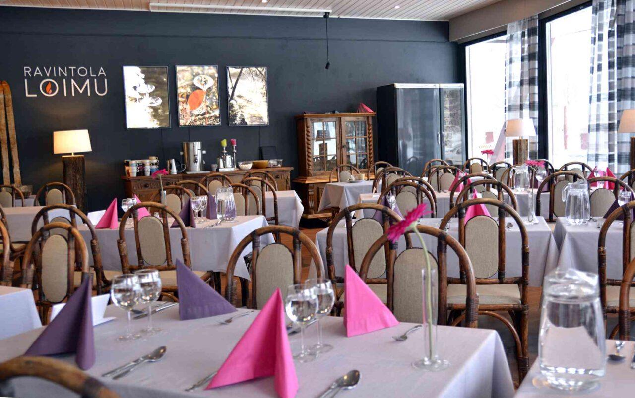 Kuvassa on valoisa ravintolasali, jossa on pöytiä katettuna valkoisilla pöytäliinoilla. Jokaisessa pöydässä on viinilasit, vesikannu sekä pinkkejä ja liloja lautasliinoja taiteltuina kolmioksi. Pöydillä on maljakoissa vaaleanpunaiset gerberat. Tuolit ovat puisia, ja niissä on vaaleat pehmusteet. Tilan takaosassa on tarjoilupöytä, jonka päällä on kahviautomaatti ja vesikannu sekä värikkäitä kynttilöitä. Seinällä lukee teksti “RAVINTOLA LOIMU”. Suuret ikkunat päästävät tilaan runsaasti luonnonvaloa, ja ikkunoiden edessä on harmaa-valkoruutuiset verhot. Yleisilme on moderni, lämmin ja kutsuva.