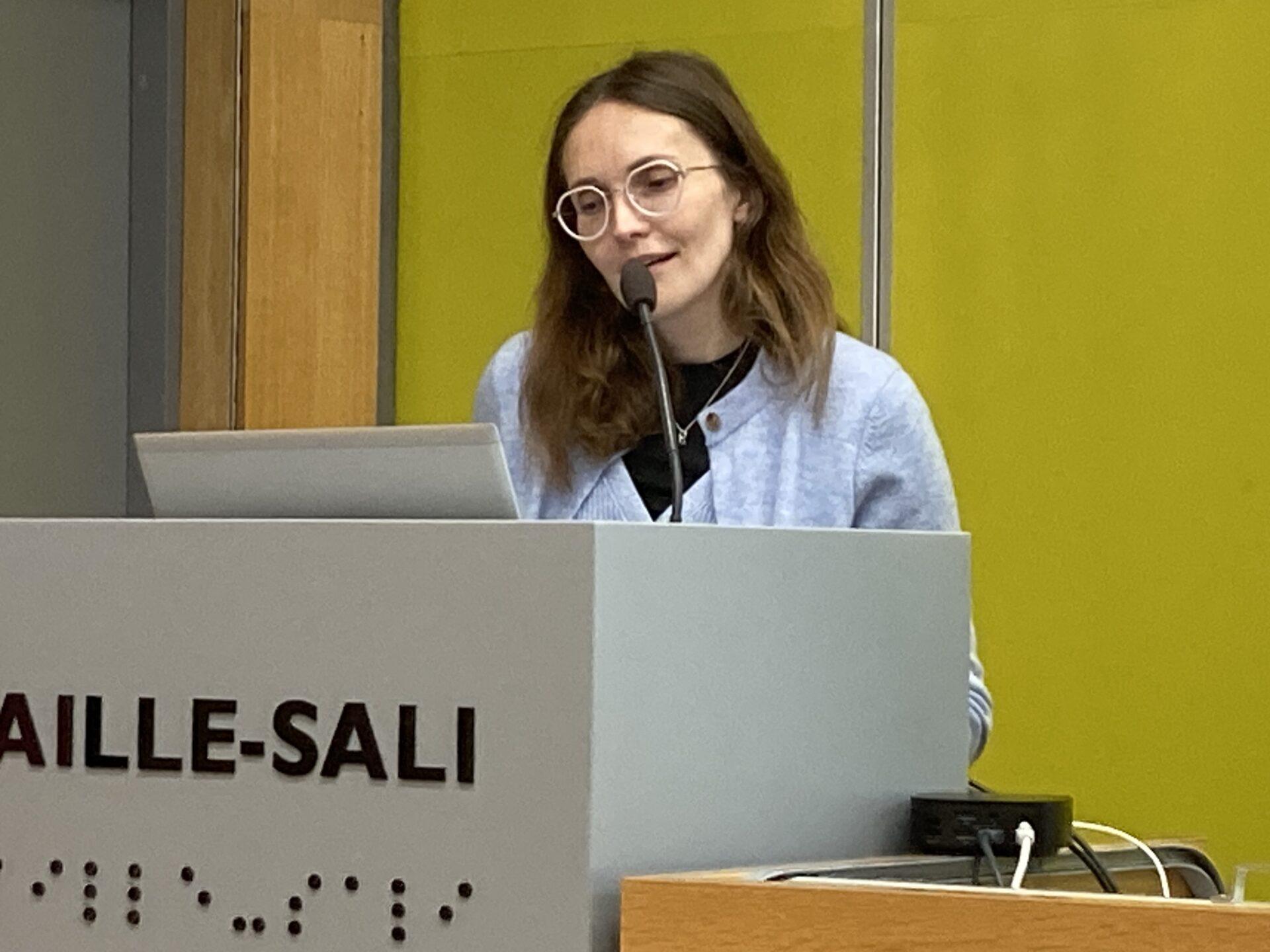 Kuvassa nainen seisoo puhujakorokkeen takana. Hänellä on vaaleat, hieman laineikkaat hiukset ja pyöreät silmälasit. Hän käyttää vaaleansinistä neuletta ja mustaa paitaa. Hän puhuu mikrofoniin ja hymyilee kevyesti. Puhujakorokkeessa on teksti BRAILLE-SALI sekä pistekirjoitusta.