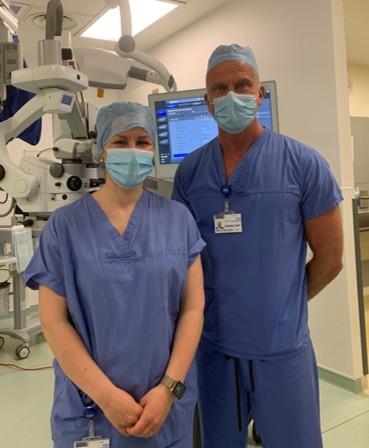 Nainen ja mies seisovat lyhythihaisissa sinisissä työasuissaan maskit kasvoilla ja päässä hiukset peittävä kertakäyttömyssy. Molemmilla on nimikyltti, jonka mies on ripustanut kaula-aukkoon, nainen lantiolleen taskun suulle. Taustalsla näkyy suurikokoinen valkoinen OCT-laite, jonka avulla seurataan reaaliajassa verkkokalvoa leikkauksen aikana. Seinällä on monitorin ruutu, jolla näkyy tekstiä ja graafinen kuva.
