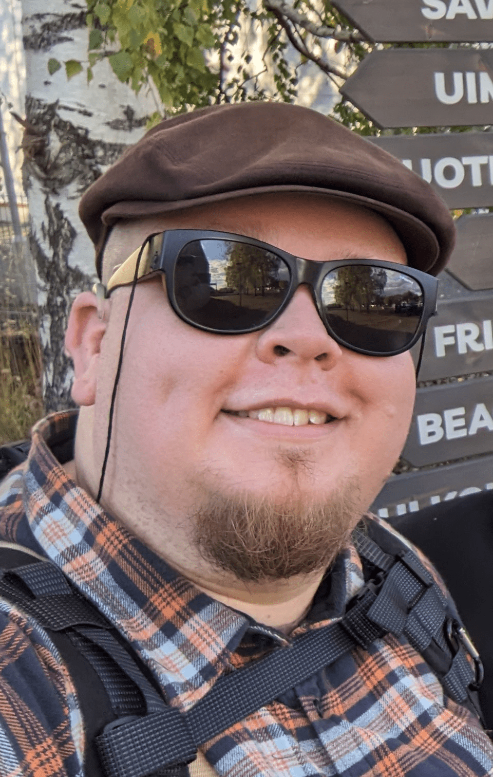 Lähikuva hymyilevästä miehestä, jolla on tummat aurinkolasit, ruskea lippalakki ja parta. Yllään hänellä on ruudullinen paita ja olalla reppu. Taustalla näkyy koivupuu sekä ruskeita opastekylttejä, joissa on valkoista tekstiä. Kyltit ohjaavat mm. rannalle ja uimarannalle. Sää näyttää aurinkoiselta.