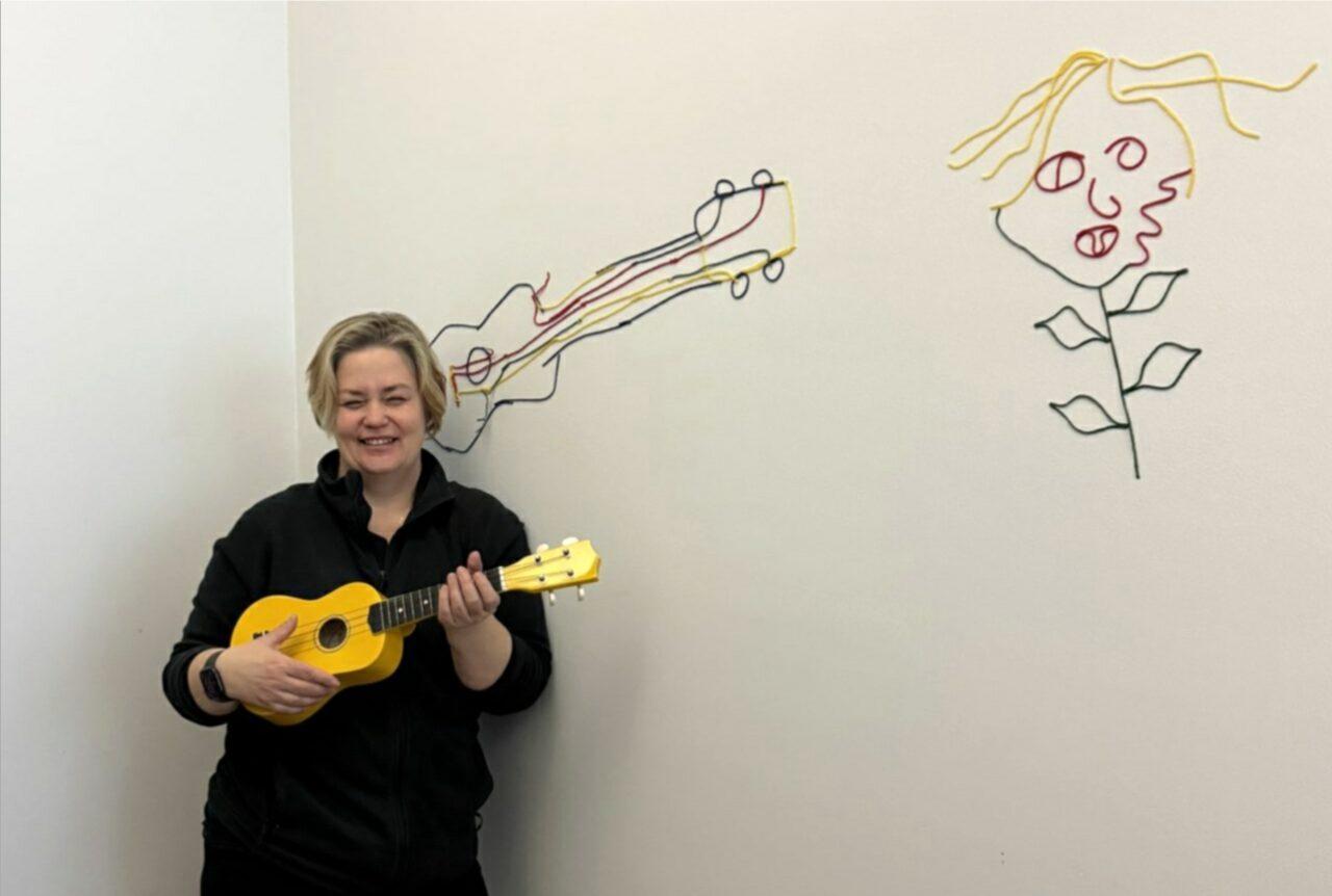 Vaaleaa seinää vasten seisoo hymyilevä nainen kädessään ukulele, joka muistuttaa pientä kitaraa. Naisella on lyhyet, vaaleat hiukset ja mustat vaatteet. Seinään on tehty langalla viivapiirroksia. Oikealla on kukka, jolla on kasvot ja liehuvat hiukset sekä varsi, jossa on neljä lehteä. Naisen pään takaa näkyy tyylitelty piirros kitarasta.