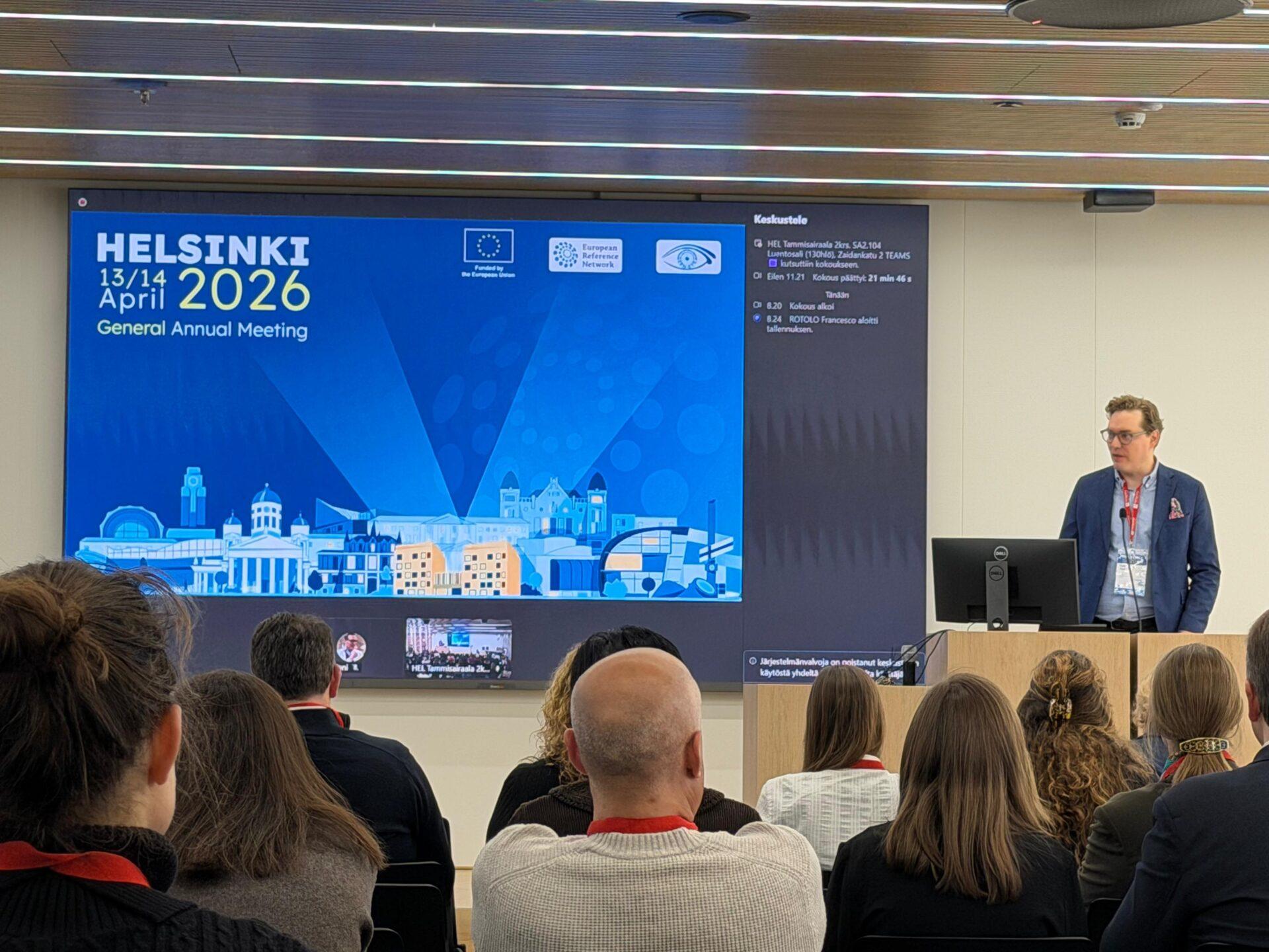 Kokoushuone, jonka suurella valkokankaalla on esitysnäyttö. Etualalla selin kameraan istuu yleisöä. Puhujakorokkeen takana seisoo mies, jolla on tummansininen bleiseri, vaalea kauluspaita ja konferenssikyltti punaisessa nauhassa. Miehen edessä on tietokonenäyttö ja pöytämikrofoni. Tunnelma on muodollinen konferenssitilaisuus. Valkokankaan yläreunassa näkyy sinisellä taustalla valkoinen teksti HELSINKI 13/14 April 2026 General Annual Meeting, sen vieressä EU:n lippu ja teksti Funded by the European Union, sitten logo, jossa lukee European Reference Network, sekä silmäsymboli. Alareunassa on piirros Helsingin rakennuksista.