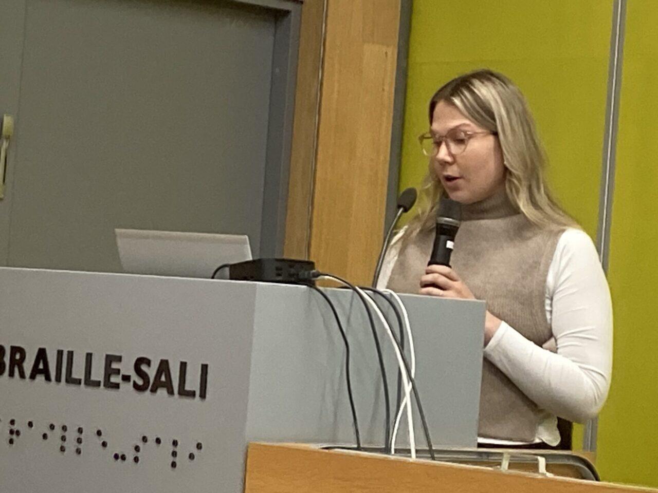 Nuori silmälasipäinen nainen seisoo puhujakorokkeen takana, jonka etuosassa lukee koho-kirjaimin ja pistekirjoituksella BRAILLE-SALI. Naisella on vaaleat, olkapäille ulottuvat hiukset ja beige neuleliivi, jonka alla on valkoinen pitkähihainen paita. Nainen puhuu ja pitää va-semmassa kädessään mustaa mikrofonia. Vaaleanharmaan puhujapöydän takaa näkyy kan-nettavan tietokoneen avattu kansi. Puhujakorokkeella on pieni musta rasia, josta roikkuu mustia ja valkoisia johtoja reunan yli. Taustalla on puunväristä pystypaneelia ja harmaa ovi.